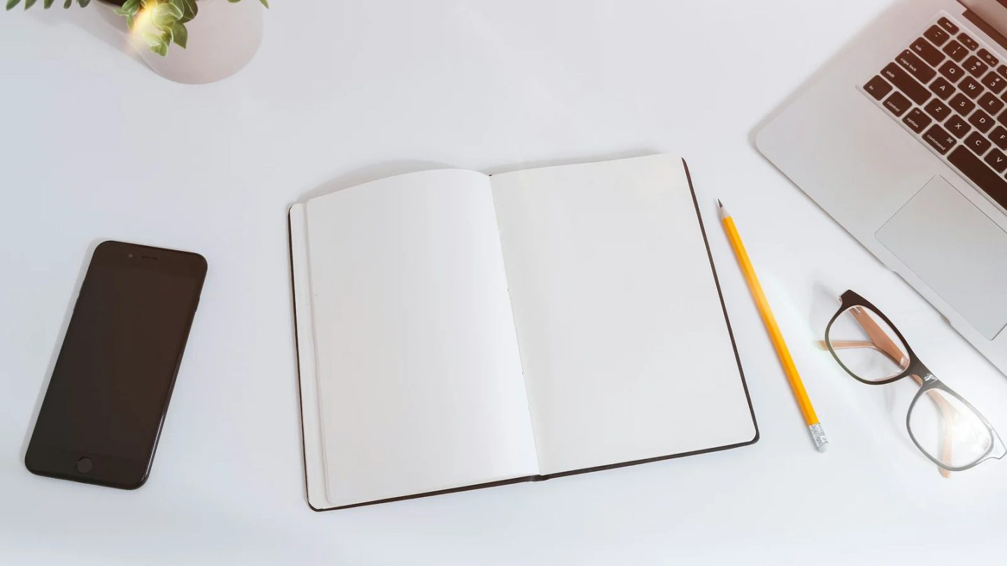 Cuaderno abierto y lápiz junto a un portátil y un teléfono sobre una mesa blanca