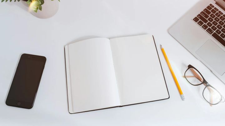 Cuaderno abierto y lápiz junto a un portátil y un teléfono sobre una mesa blanca